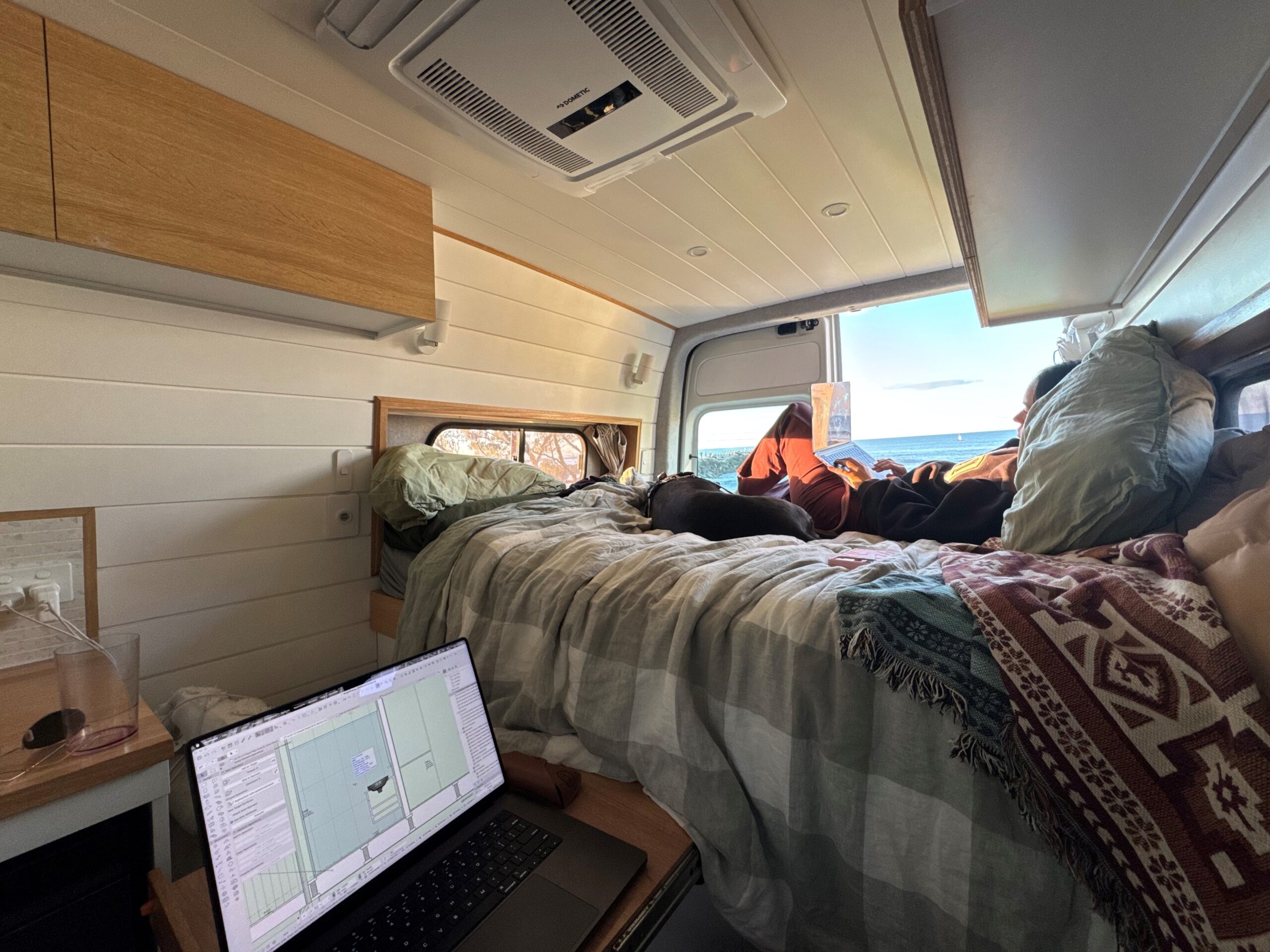 Casey drafting on a laptop in the van, with Bean on the bed and the ocean through the open door